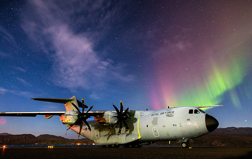 Photo: Atlas C Mk. 1 aircraft parked on an airfield in Greenland. Above it, the colours of the Aurora Borealis are in the night sky.